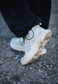 Witte trailrunningschoen met een gestructureerde zool die op rotsachtige grond stapt, gedragen met een witte sok en zwarte broek.