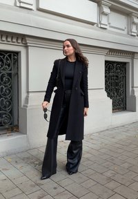 Schwarzer Wollmantel mit einem doppellagigen Design, kombiniert mit einem enganliegenden schwarzen Oberteil und weit geschnittenen Lederhosen. Sanfte Texturen; minimaler Schmuck.