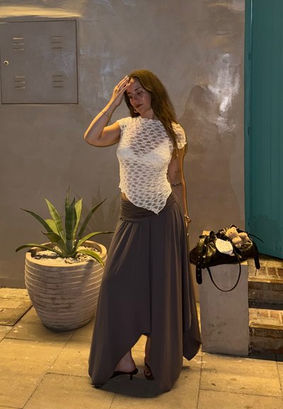 Top blanco texturizado con mangas cortas y dobladillo asimétrico, combinado con una falda gris fluida. Bolso negro con acentos decorativos. Planta en maceta cercana.