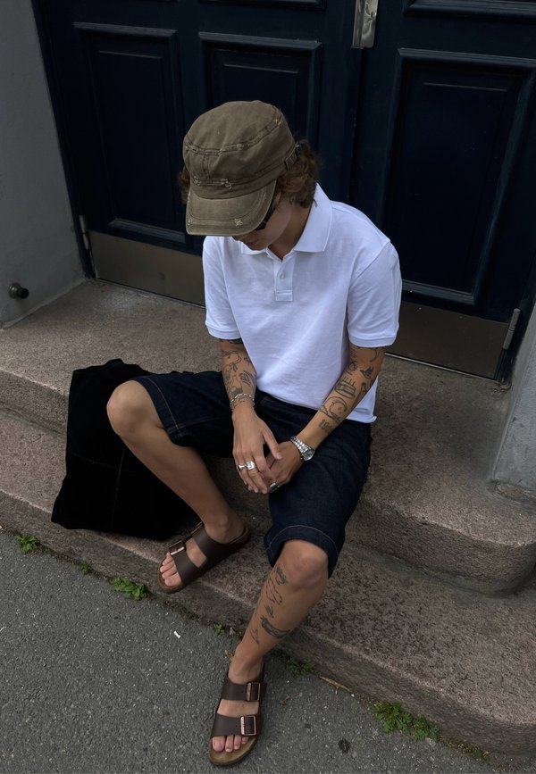 Camisa polo branca, calções de ganga escuros, sandálias castanhas, relógio prateado, múltiplos anéis, múltiplas tatuagens, usando um boné verde, sentado em degraus de pedra.