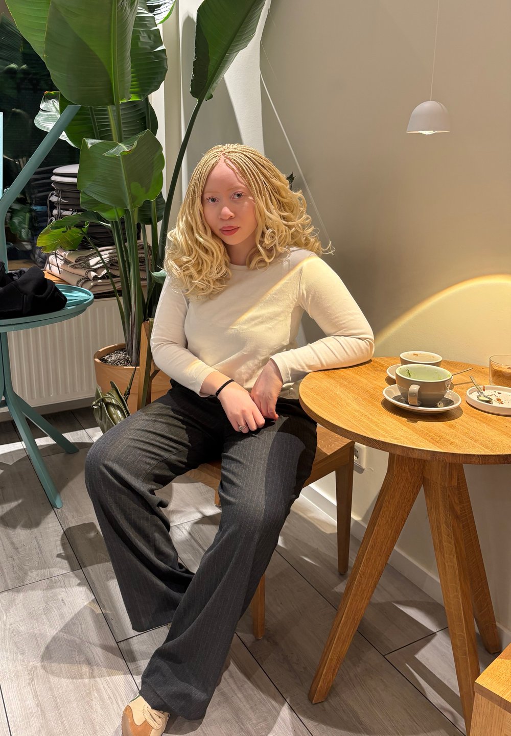 Une personne est assise sur une chaise en bois à une table ronde. Elle porte une chemise manches longues beige clair et un pantalon rayé foncé. Une plante en pot est visible à proximité.