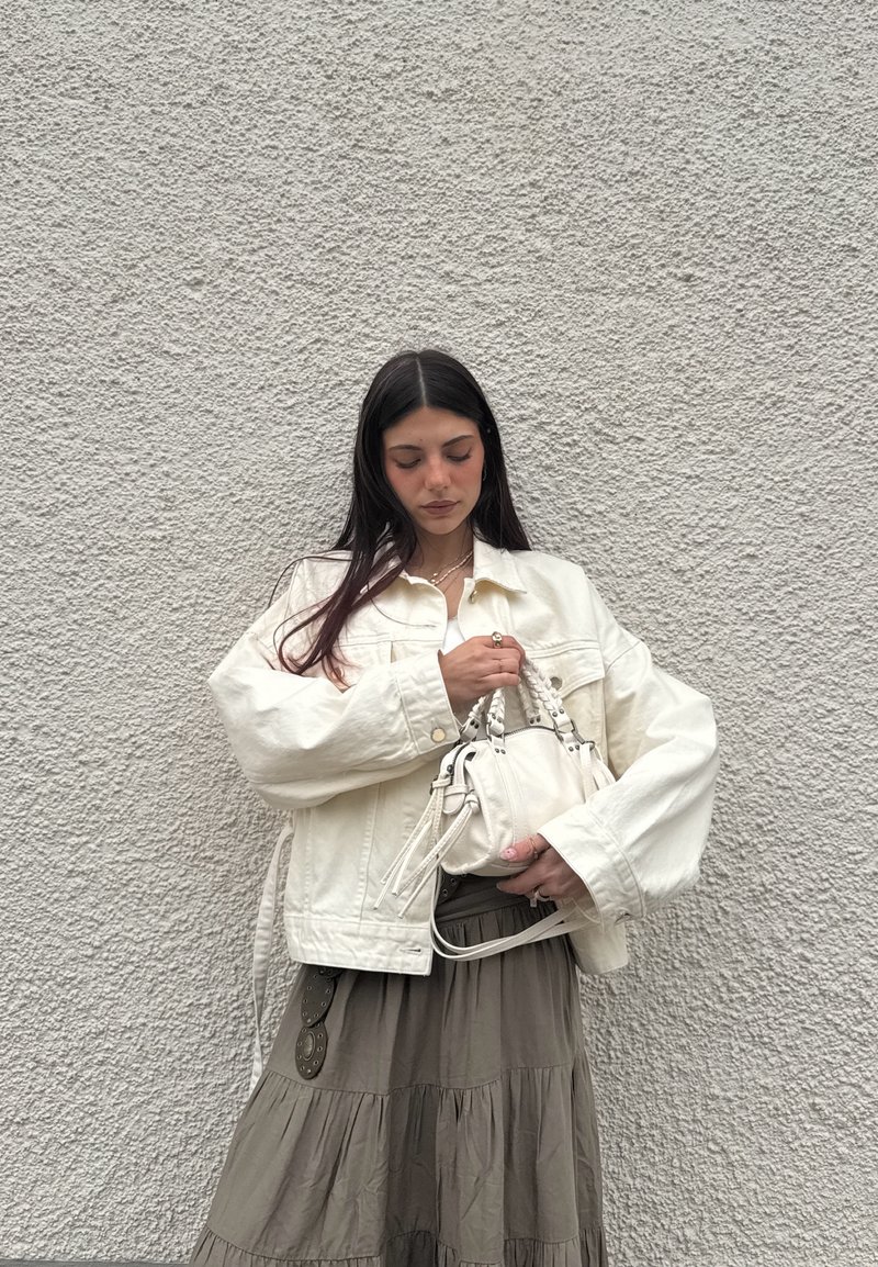 Femme aux longs cheveux foncés portant une veste blanche et une jupe grise, tenant un sac à main blanc devant un mur texturé gris clair.