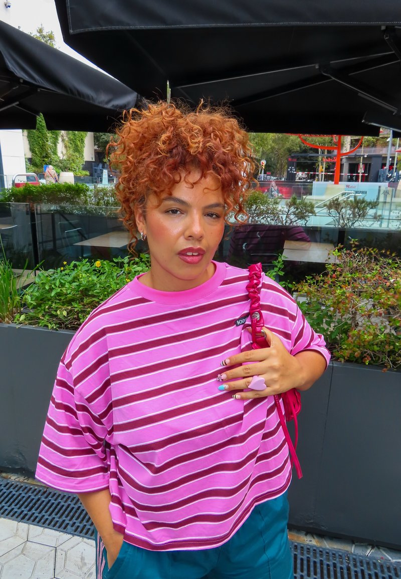 Personne aux cheveux roux bouclés portant une chemise rose à rayures et un pantalon turquoise se tient dehors près des plantes, tenant une sangle de sac rouge sur l'épaule.