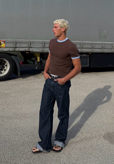 Camiseta marrón de manga corta con ribete azul claro, jeans azul oscuro y chanclas negras. De pie al aire libre junto a una camioneta.