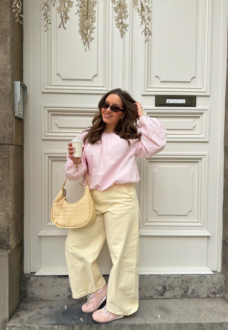 Γυναίκα στέκεται μπροστά σε λευκή πόρτα κρατώντας κούπα καφέ, φοράει ροζ μπλούζα, κρεμ παντελόνι φαρδιά, ροζ αθλητικά παπούτσια, γυαλιά ηλίου και μπεζ τσάντα.