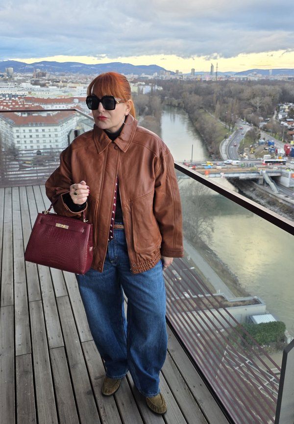 Femme aux cheveux roux portant une veste en cuir marron et des lunettes de soleil, tenant un sac à main rouge, debout sur un balcon donnant sur une rivière et le paysage urbain.