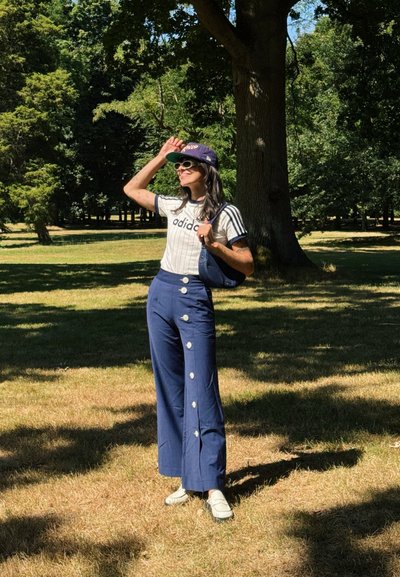 Camiseta a rayas blancas y marinas, pantalones de pierna ancha azul con grandes botones blancos, zapatos blancos, gorra negra y gafas de sol en un parque cubierto de hierba.