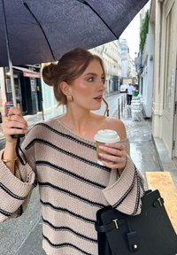 Dicker Strickpullover in Beige mit schwarzen Streifen, hält einen Kaffeebecher und einen Regenschirm, dazu eine schwarze Tasche mit goldenen Akzenten.