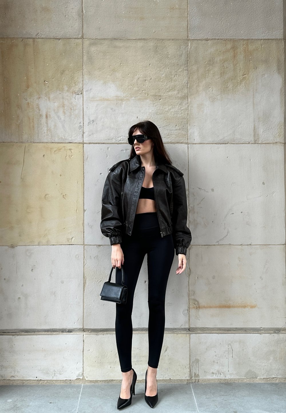 Mujer vestida con chaqueta de cuero negra, top corto negro, leggings, tacones altos, gafas de sol, sosteniendo un pequeño bolso negro, de pie contra una pared de azulejos marrones.