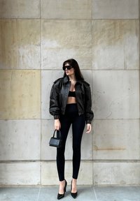 Mujer vestida con chaqueta de cuero negra, top corto negro, leggings, tacones altos, gafas de sol, sosteniendo un pequeño bolso negro, de pie contra una pared de azulejos marrones.