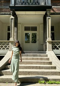 Jasnozielona, zwiewna maxi sukienka z off-shoulder, uszyta z miękkiego materiału, o luźnym kroju, noszona przez postać schodzącą po kamiennych schodach.