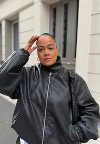 Jaqueta de couro preta com gola alta, fecho de prata e punhos elásticos. Textura suave com bolsos laterais e uma alça de ombro trançada.