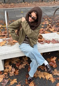 Le mannequin porte une écharpe en tricot marron et une veste vert olive sur un haut rayé. Un jean bleu clair usé et des mocassins marron foncé complètent le look.