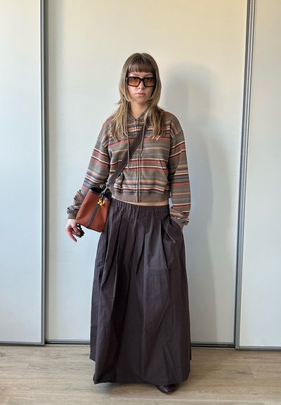 Persona joven con chaqueta con cremallera a rayas, falda larga oscura, gafas de sol y llevando un bolso marrón de bandolera, de pie en el interior sobre suelo de madera.