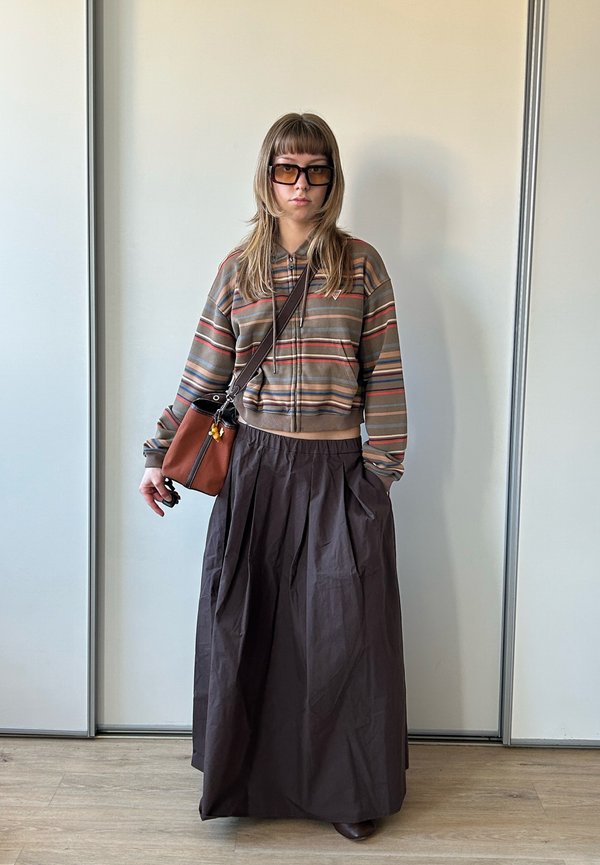 Jong persoon draagt een gestreept jack met rits, lange donkere rok, zonnebril en draagt een bruine crossbodytas, staat binnen op een houten vloer.