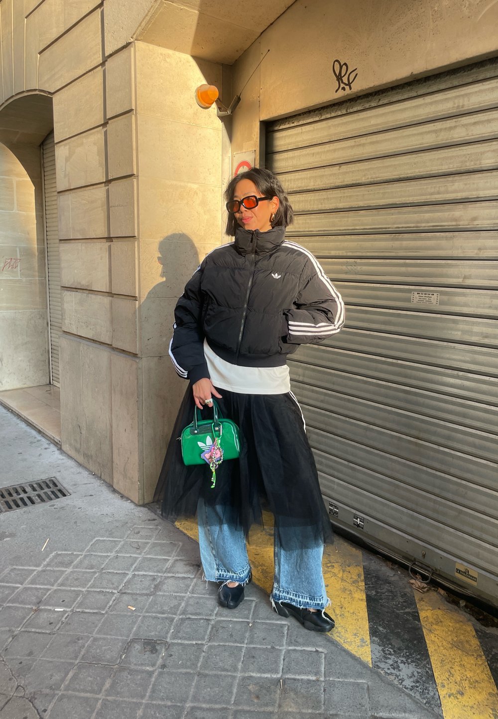 Femme en doudoune noire, jupe en tulle noir portée sur un jean, tenant un sac à main vert, debout contre un mur beige et une porte métallique fermée.