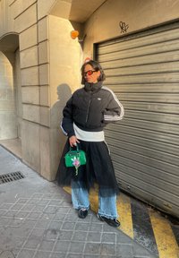 Femme en doudoune noire, jupe en tulle noir portée sur un jean, tenant un sac à main vert, debout contre un mur beige et une porte métallique fermée.