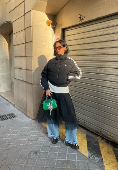 Mujer con chaqueta acolchada negra, falda de tul negra sobre jeans, sosteniendo un bolso verde, de pie contra una pared beige y una persiana metálica cerrada.