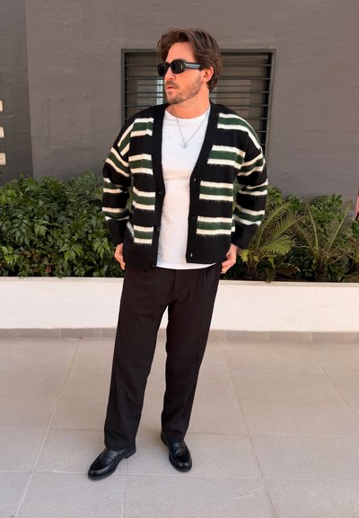 Hombre con cárdigan de rayas blancas y negras, camisa blanca, pantalones negros y gafas de sol, de pie sobre suelo de baldosas cerca de plantas y pared gris.