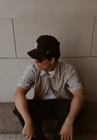 Giovane uomo che indossa una camicia a maniche corte a scacchi, occhiali da sole e cappellino nero, seduto contro un muro di piastrelle beige, che guarda di lato.