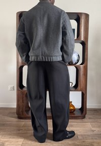 Personne portant une veste grise et un pantalon large noir, debout les mains dans les poches devant une étagère en bois avec des objets décoratifs.