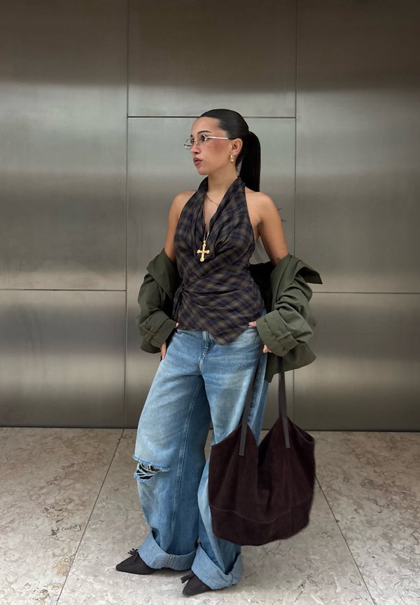 Une femme portant des lunettes pose avec un haut à carreaux noué derrière le cou, un jean ample et une veste, en tenant un grand sac sombre devant un mur métallique.