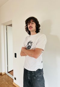 T-shirt branca com um desenho gráfico preto, jeans escuros rasgados, encostado a uma parede clara com chão de madeira e moldura da porta visíveis.