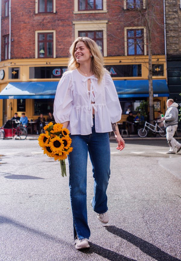 Top blanco con mangas abullonadas y detalle de lazo, vaqueros azul claro y zapatillas blancas. Sosteniendo un ramo de girasoles, caminando por una calle.