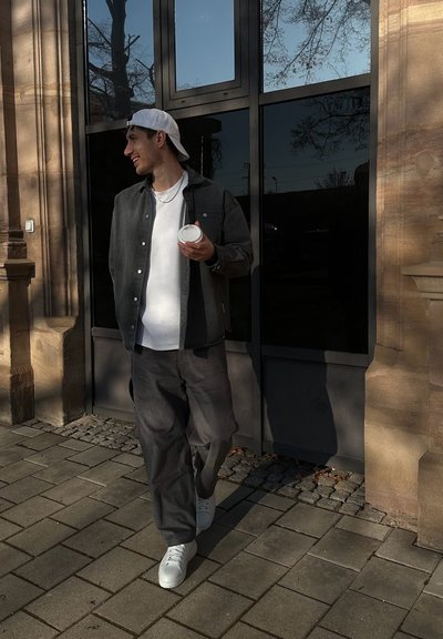 Hombre con una chaqueta de botón negra y gris sobre una camisa blanca, sosteniendo una taza, vistiendo pantalones grises y zapatillas blancas, caminando junto a puertas de cristal.