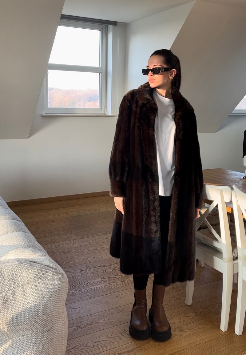Μακρύ παλτό από τεχνητό γούνινο ύφασμα σε σκούρο καφέ, με φαρδύ γιακά, φορεμένο πάνω από λευκή μπλούζα, συνδυασμένο με μαύρες κολάν και χοντρά καφέ μποτάκια.