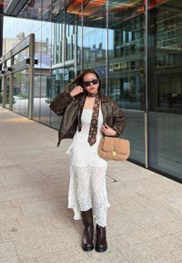 Bruine leren oversized bomberjack, witte kantjurk met asymmetrische zoom, bruin kniehoge laarzen, gedessineerde sjaal en gestructureerde lichtbruine handtas.