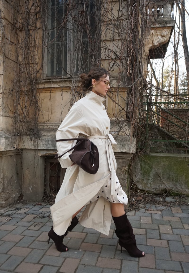 Žena v béžovom trenčkote, šatách s bodkami a čiernych vysokých podpätkoch kráča okolo starej budovy pokrytej brezlistými viničmi.