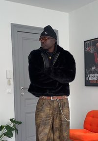 Homme portant un bonnet noir, des lunettes de soleil marron, une veste en fourrure noire et un jean marron usé, debout les bras croisés dans une pièce avec une affiche de basket-ball et une chaise orange.