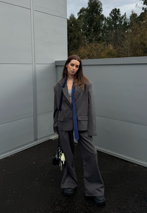 Woman wearing an oversized grey suit and blue tie, standing outdoors near grey walls with trees in the background, holding a black handbag.