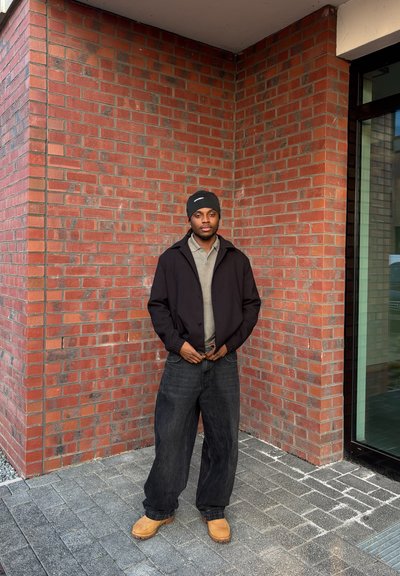 Chaqueta negra sobre una camiseta gris, vaqueros anchos oscuros y botas color tan. Situado frente a una pared de ladrillo rojo texturizada con una ventana de cristal.