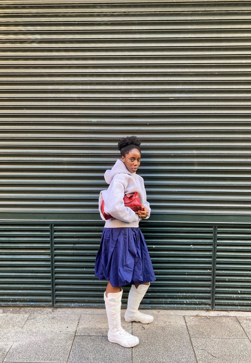 Hanorac gri cu glugă, fustă plisată bleumarin, cizme albe până la genunchi și o geantă roșie; modelul stă în fața unui fundal verde ondulat.