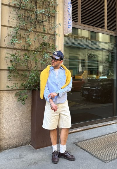 Hombre con camisa azul, suéter amarillo sobre los hombros, pantalones cortos beige, calcetines blancos, zapatos oscuros, gorra negra y gafas, de pie en una calle de la ciudad cerca de un restaurante.