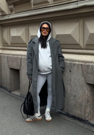 Mujer con sudadera con capucha gris, abrigo largo gris, mallas, zapatillas deportivas, gafas de sol grandes, de pie en la acera junto a una pared de piedra, sosteniendo una bolsa negra.
