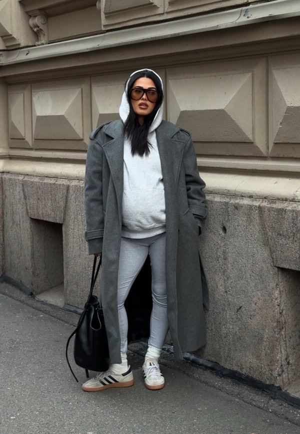Kvinde i grå hættetrøje, lang grå frakke, leggings, sneakers, store solbriller, står på fortovet mod en stenvæg, holder en sort taske.