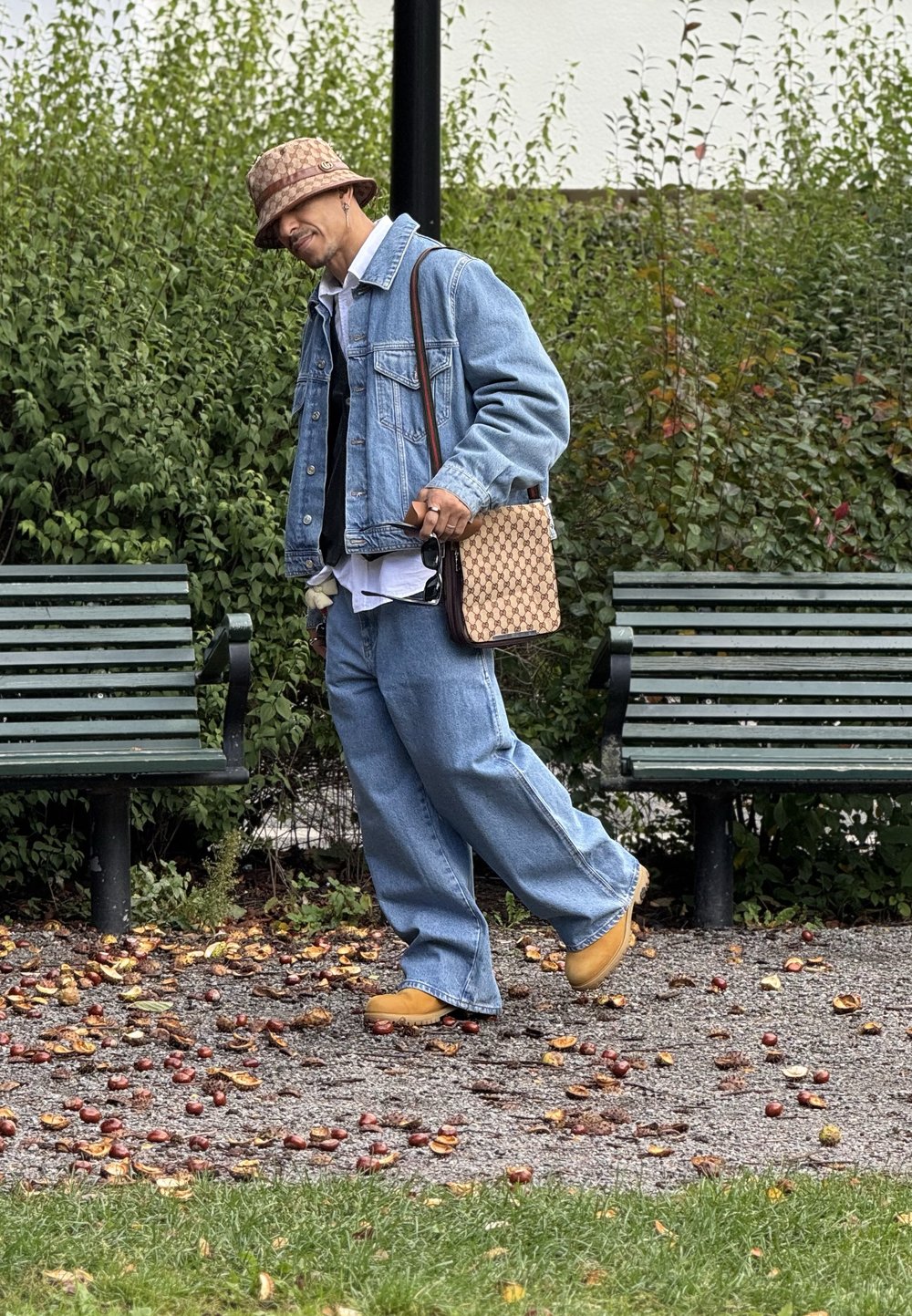 Jeansjacke über einem weißen Hemd, lockere blaue Jeans, gelbe Schuhe und eine gemusterte braune Tasche. Hintergrund aus grüner Vegetation und Bänken.