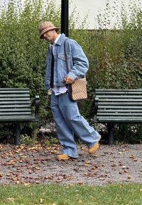 Jeansjacke über einem weißen Hemd, lockere blaue Jeans, gelbe Schuhe und eine gemusterte braune Tasche. Hintergrund aus grüner Vegetation und Bänken.