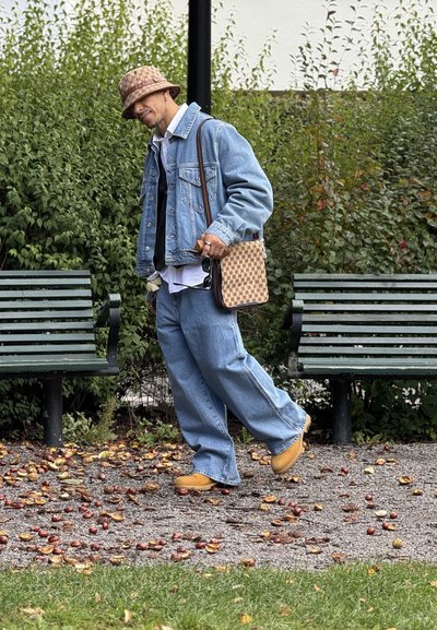 Chaqueta de mezclilla sobre una camisa blanca, jeans azules holgados, calzado amarillo y bolso marrón con estampado. Fondo de follaje verde y bancos.