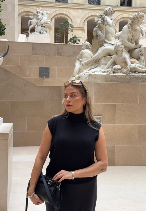Femme en haut noir sans manches et jupe, debout à l'intérieur d'un musée près de sculptures classiques en marbre blanc sur des murs en pierre beige.
