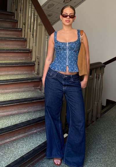 Top de corsé de mezclilla con estampado floral, pantalones acampanados de tiro alto en azul oscuro y bolso de ante marrón; gafas de sol negras completan el look.