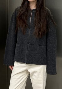 Veste en laine grise à encolure ronde, fermetures à boutons à bascule, manches longues et deux poches avant. Associée à un pantalon blanc.