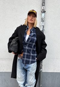 Femme blonde portant une casquette marron, un manteau noir, une chemise à carreaux bleue, un débardeur blanc, un jean clair déchiré, tenant un sac noir, debout contre un mur avec un poteau couvert d'autocollants.