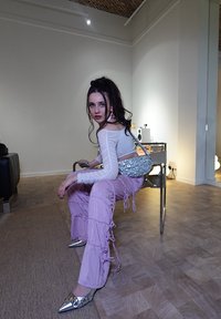 Ασημένια τσαντάκι γοναρίσματος με παγιέτες, λευκή μπλούζα με μακριά μανίκια, ροζ παντελόνι με φραμπαλά και μεταλλικές μυτερές γόβες με διακοσμητικά στοιχεία. Εσωτερικός χώρος με ξύλινο δάπεδο.