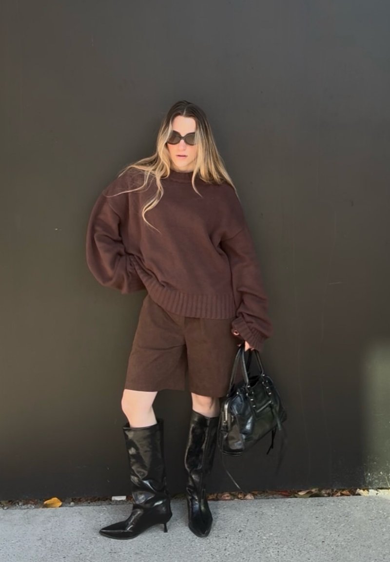 Γυναίκα που φοράει υπερμεγέθη καφέ πουλόβερ και σορτς, μαύρες μπότες μέχρι το γόνατο, γυαλιά ηλίου, κρατώντας μαύρη τσάντα, στέκεται μπροστά σε σκοτεινό τοίχο.