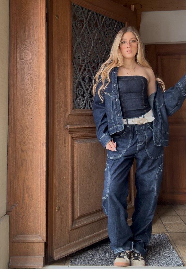 Ung kvinne med langt blondt hår iført denimjakke og bukser, stroppeløs topp, hvitt belte og briller, står ved en tredør.