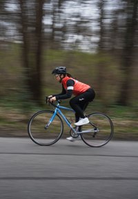 Ciclista con un gilet rosso e leggings neri su una bici da strada blu, indossando un casco nero e occhiali, che pedala su un percorso asfaltato sfocato.